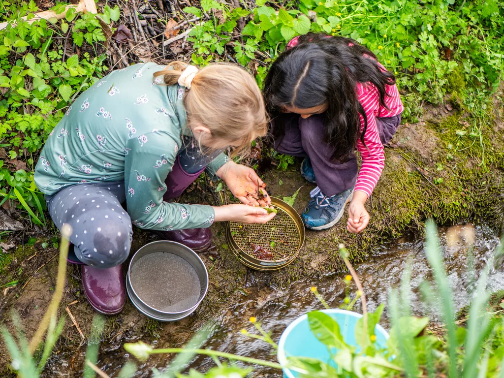 Zwei Kinder sitzen an einem Bach und untersuchen mit Sieben Steine oder Erde im Wasser.