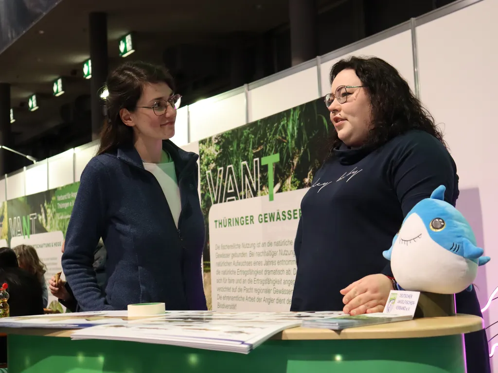 Zwei Frauen unterhalten sich an einem Messestand; auf dem Tresen liegt ein Plüschhai.