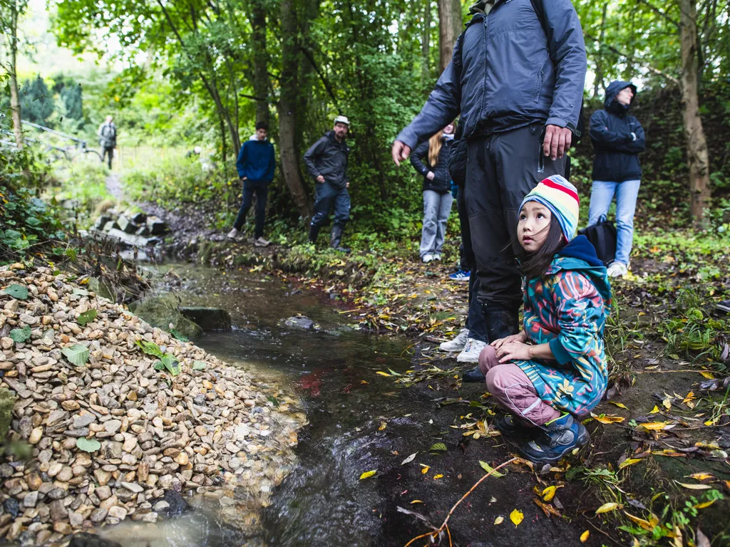 Ein Kind mit bunter Mütze hockt am Ufer eines kleinen Bachs im Wald, umgeben von mehreren Erwachsenen.