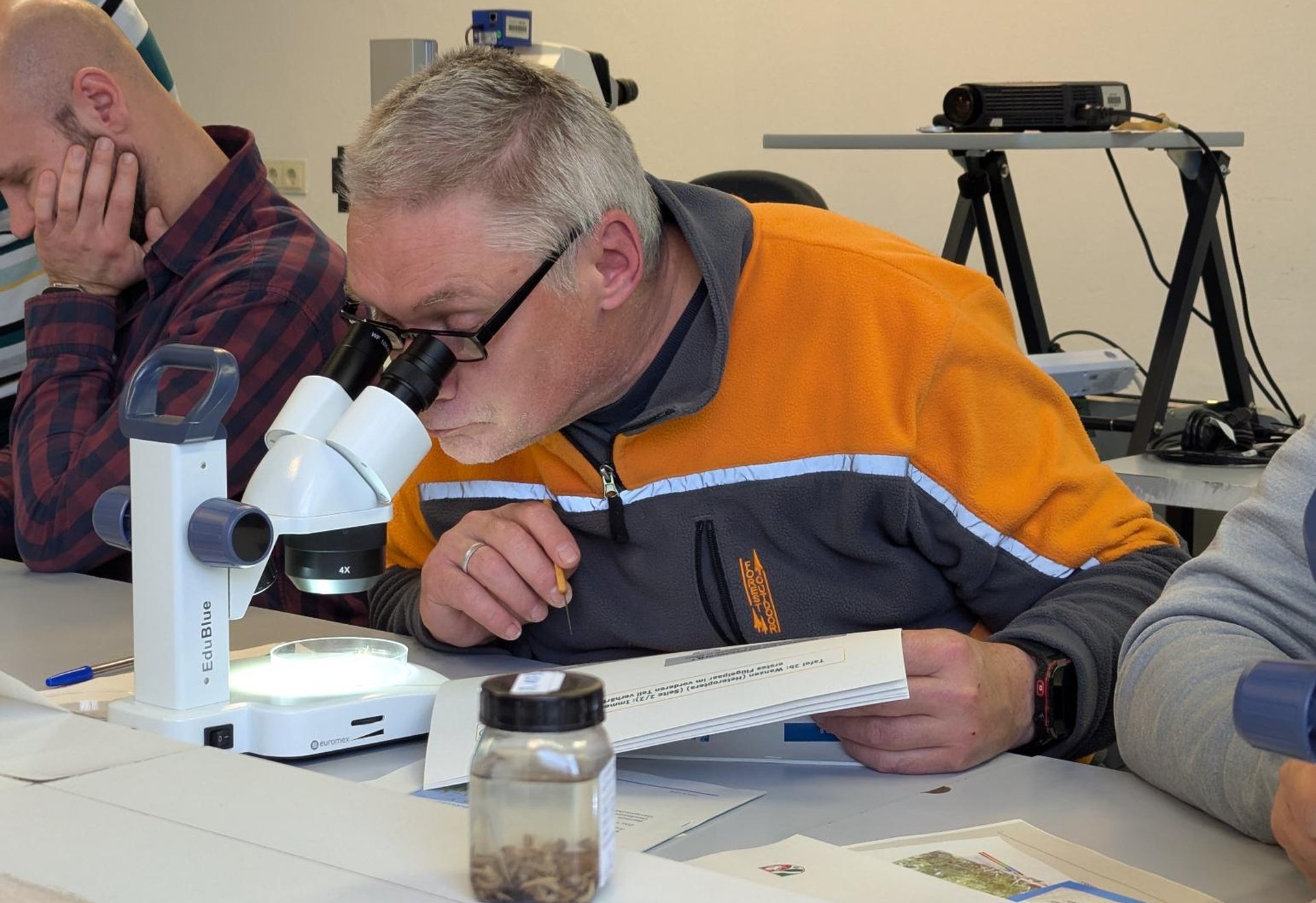 Eine Person beugt sich über ein Mikroskop und untersucht eine Probe. Auf dem Tisch liegen Unterlagen, ein Glas mit Probenmaterial und weiteres Laborzubehör.