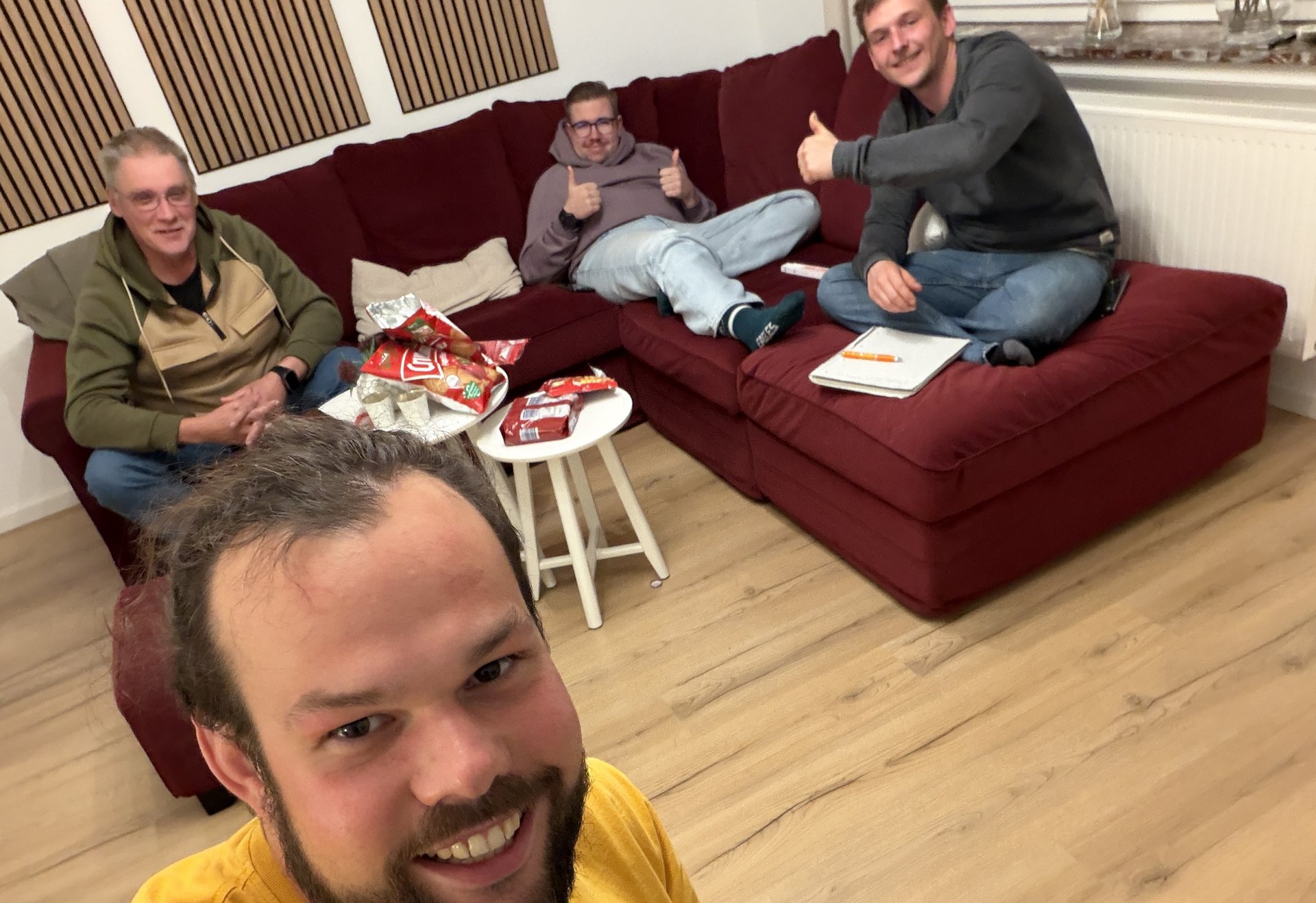 Vier Männer sitzen und lachen in einem Wohnzimmer auf einem roten Sofa, einer macht ein Selfie, auf dem Tisch liegen Snacks.