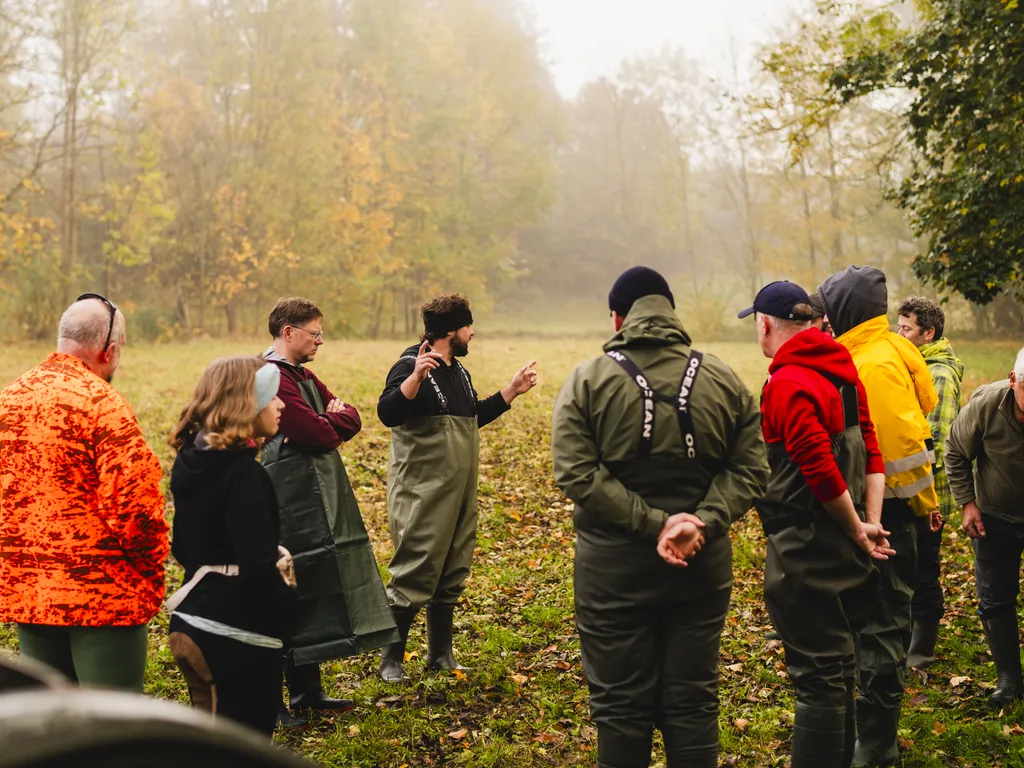 Eine Gruppe von Menschen steht im Freien auf einer Wiese im Herbst und hört einer Person in der Mitte aufmerksam zu.
