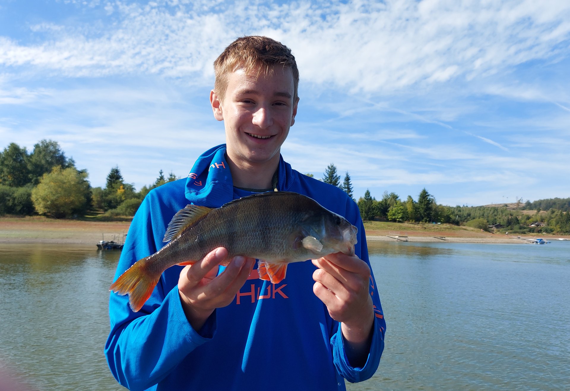 Ein junger Mann hält einen Fisch vor einem See und lächelt in die Kamera.