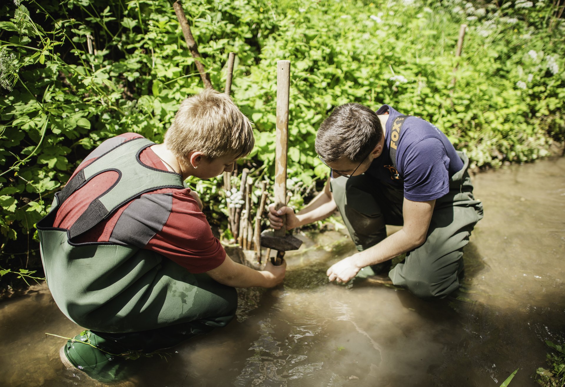 Zwei Jugendliche knien in einem flachen Bach und schlagen Holzpfähle in den Boden, um eine Uferstruktur zu befestigen.