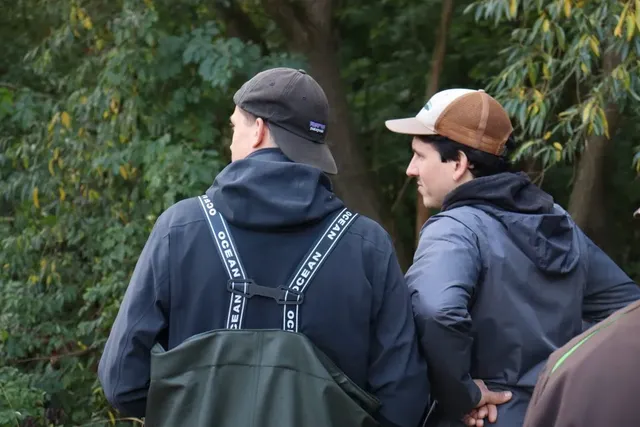 Zwei Männer mit Kappen stehen im Freien und unterhalten sich, im Hintergrund sind Bäume zu sehen.