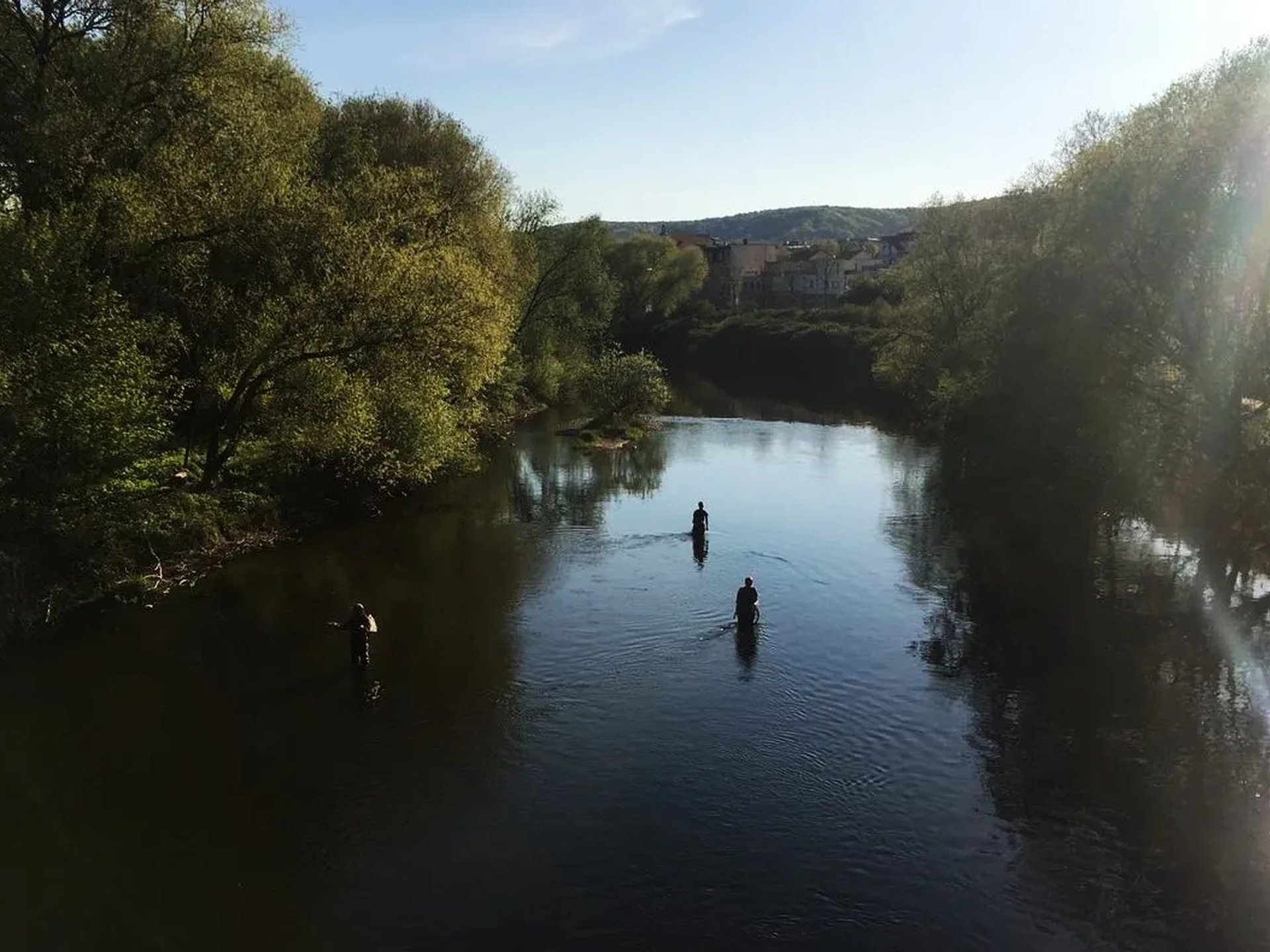 Drei Menschen stehen im Fluss und angeln, umgeben von Bäumen und Sonnenlicht.