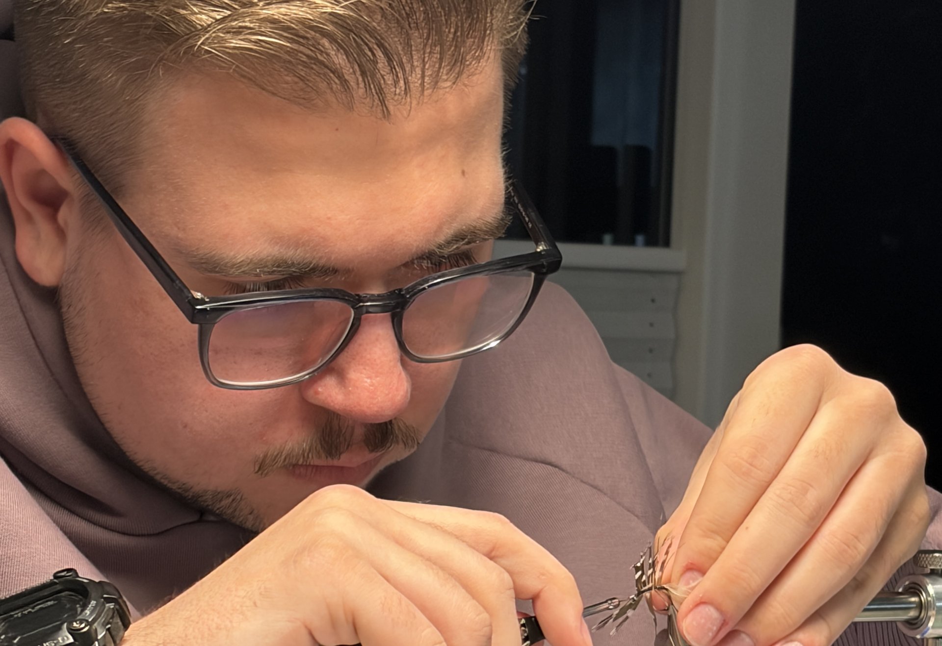 Ein Mann mit Brille und Bart knotet konzentriert eine künstliche Fliege an einem Bindestock.
