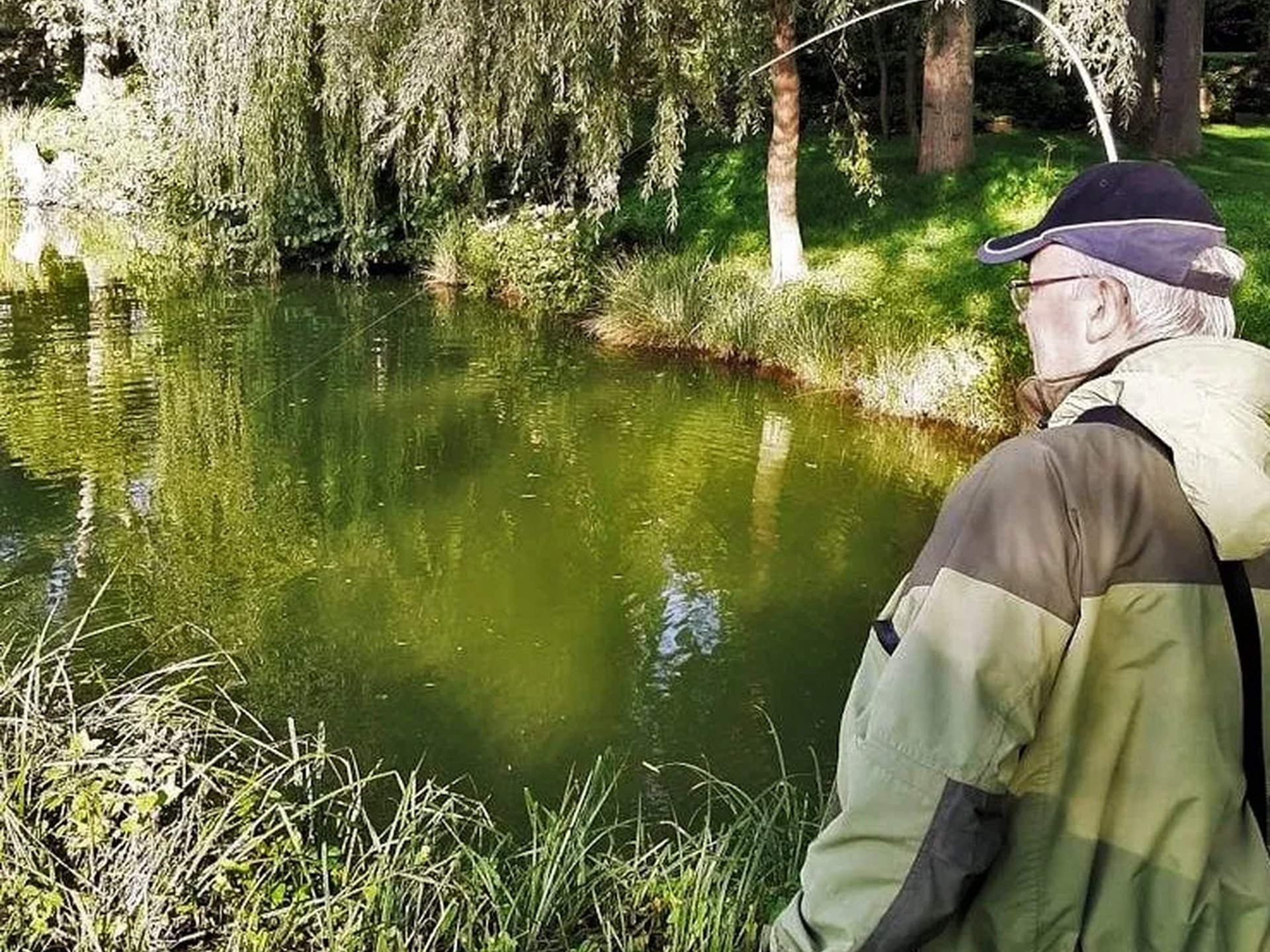 Ein Mann mit Angelkleidung steht am Ufer eines grünen Teichs und angelt.