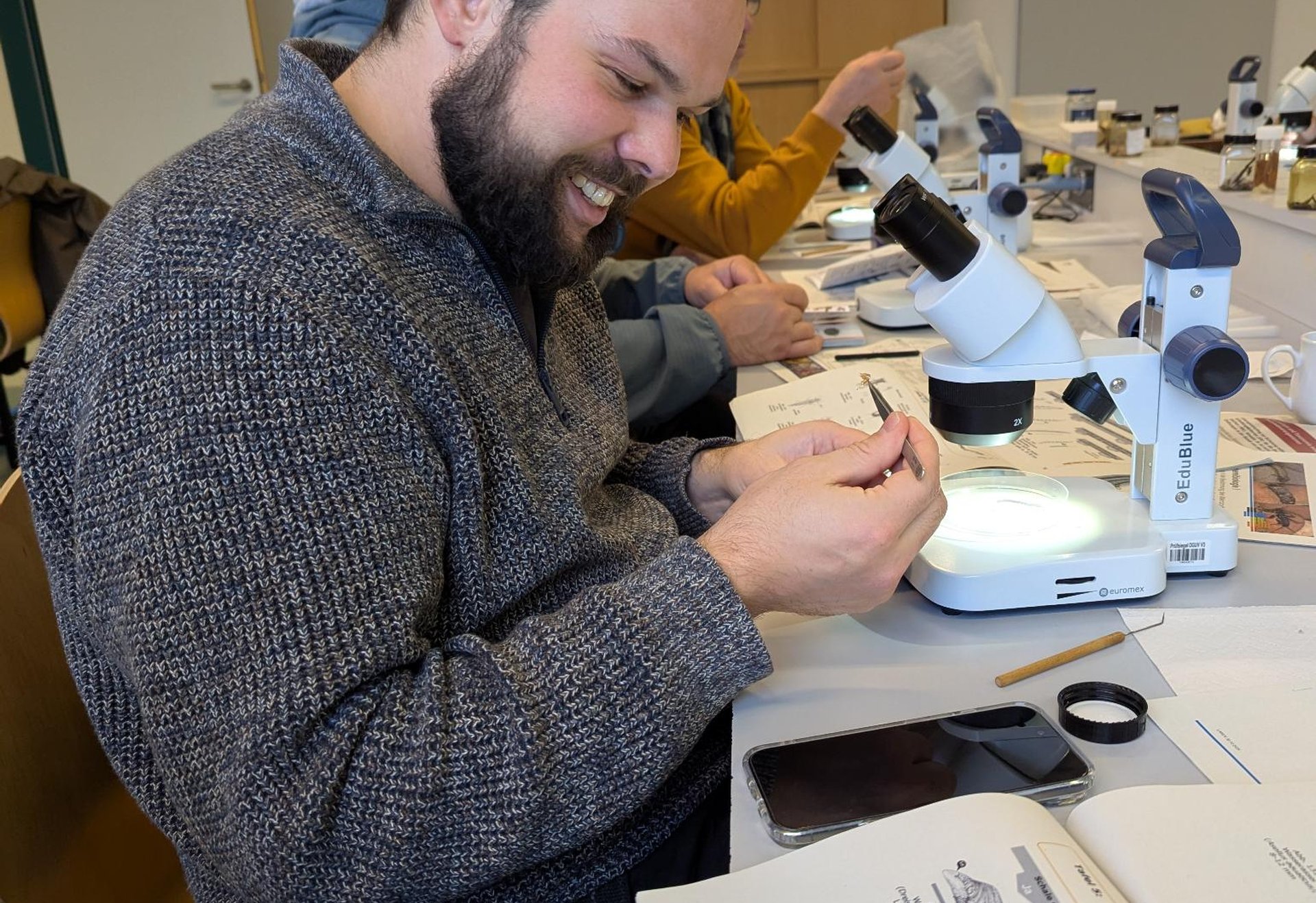 Eine Person sitzt an einem Tisch und schaut durch ein Stereomikroskop. In einer Petrischale unter dem Mikroskop liegt eine Probe. Daneben liegen Unterlagen und weitere Materialien.