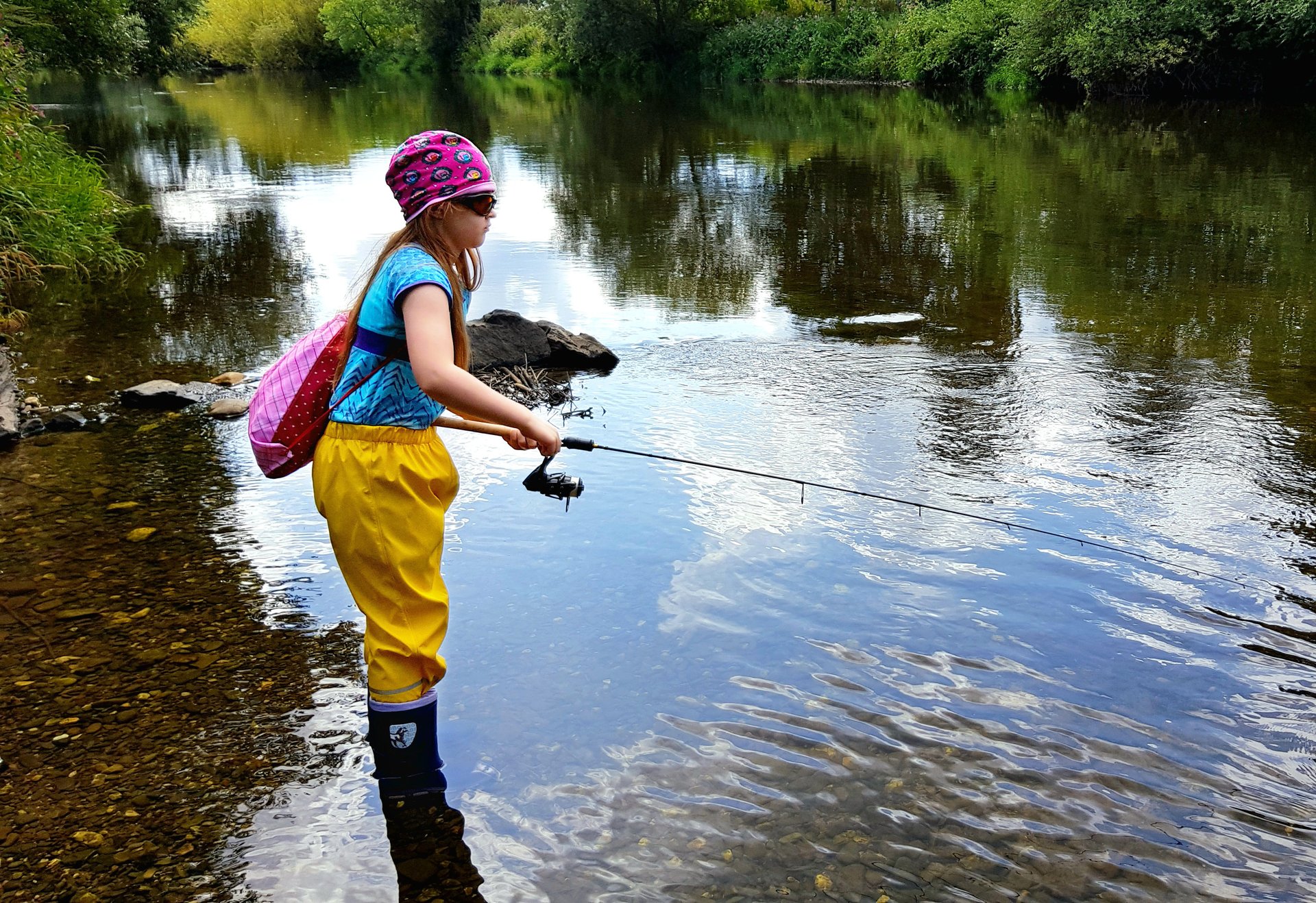 Ein Mädchen in gelber Regenhose angelt an einem ruhigen Flussufer.