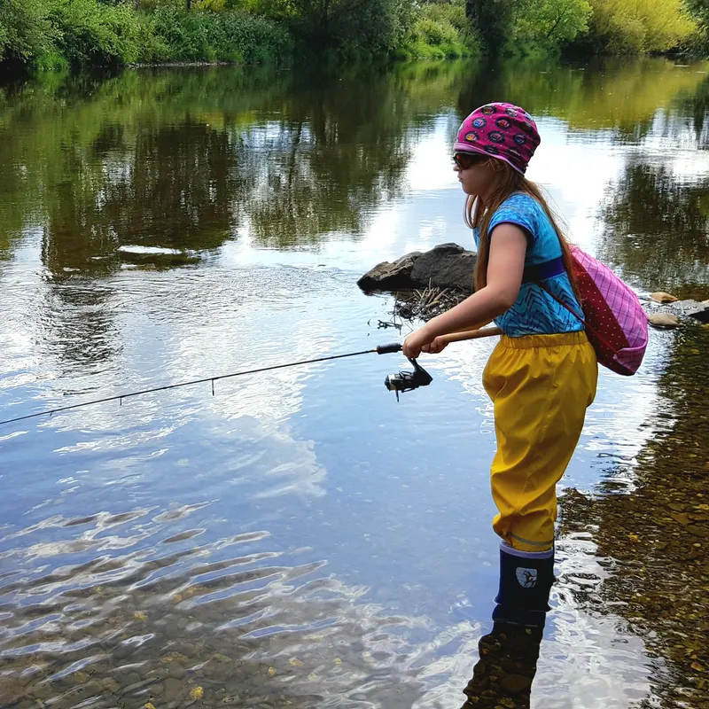 Ein Kind steht in einem flachen, klaren Fluss und angelt mit einer kleinen Angelrute. Es trägt eine bunte Mütze, eine Sonnenbrille, eine blaue Kurzarmbluse, gelbe wasserdichte Hosen und Gummistiefel. Auf dem Rücken trägt es einen pinken Rucksack. Die Umgebung ist von dichter grüner Vegetation gesäumt, und die Wasseroberfläche reflektiert den Himmel und die Bäume.