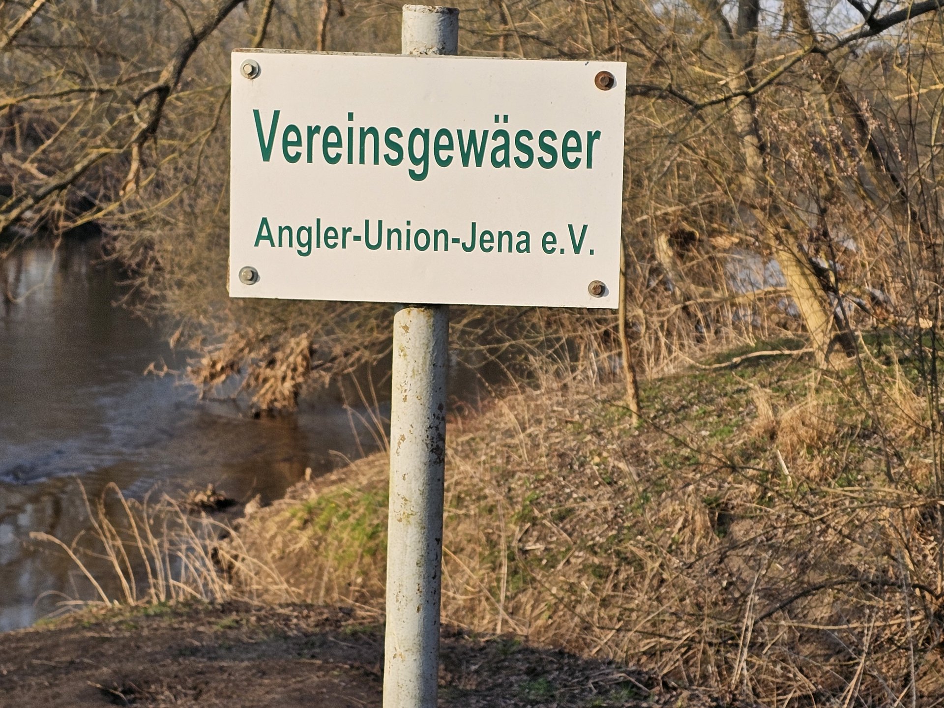Ein Schild mit der Aufschrift „Vereinsgewässer Angler-Union-Jena e.V.“ steht am Ufer eines Flusses in einer natürlichen, bewaldeten Umgebung.