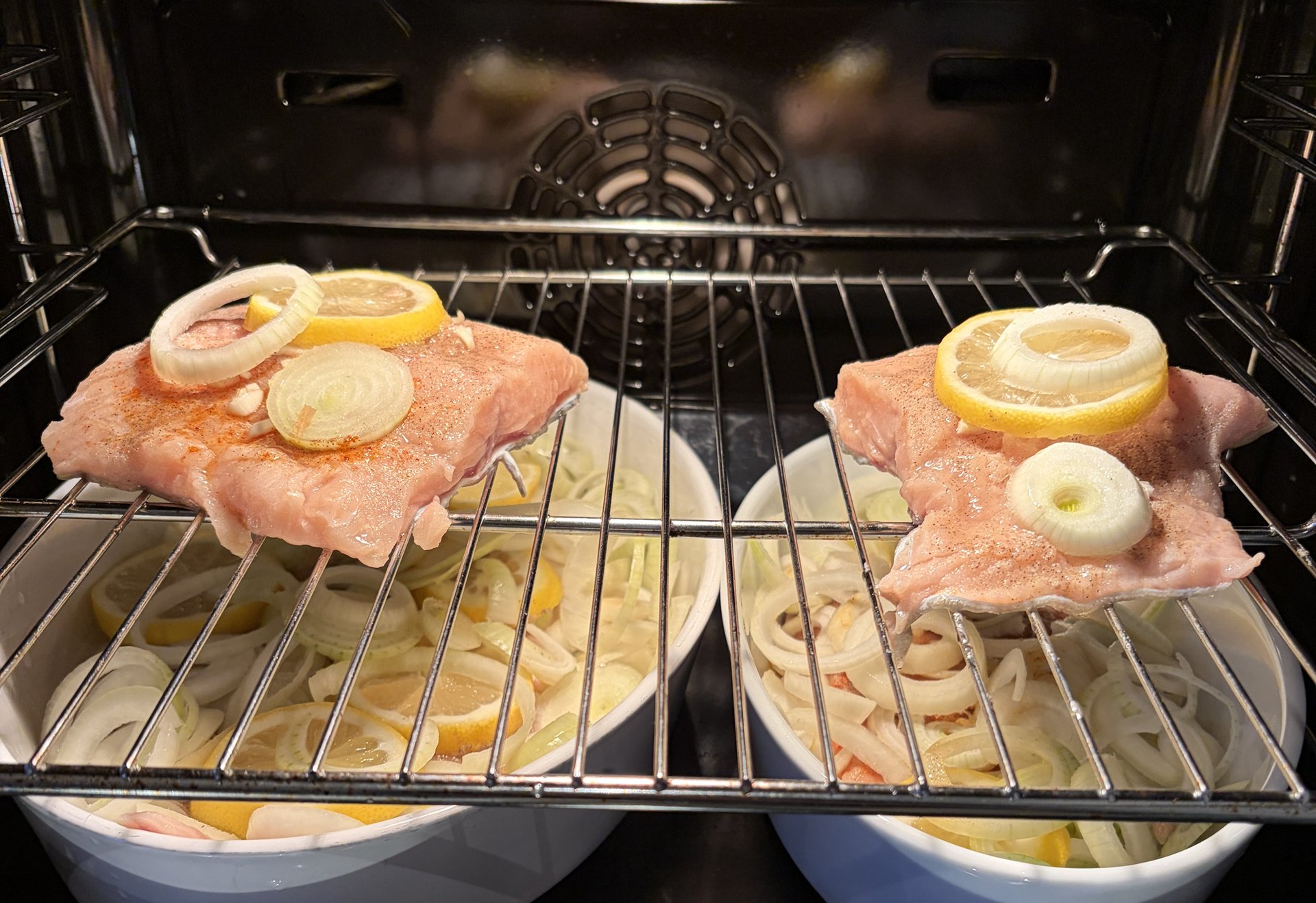 Zwei Stücke Fisch mit Zitronen- und Zwiebelscheiben liegen auf einem Rost im Ofen, darunter stehen zwei Auflaufformen mit Gemüse.