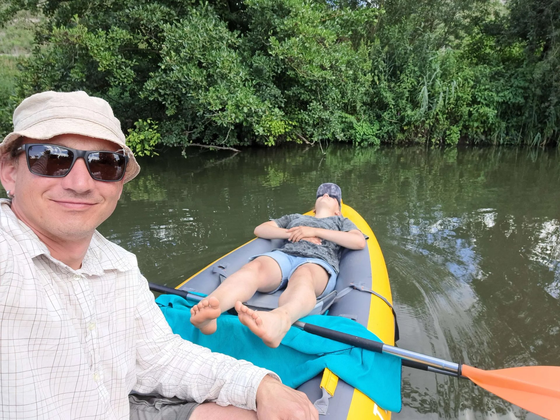 Zwei Personen im Kajak; eine sitzt lächelnd vorne mit Hut und Sonnenbrille, die andere liegt entspannt auf dem Rücken und sonnt sich.