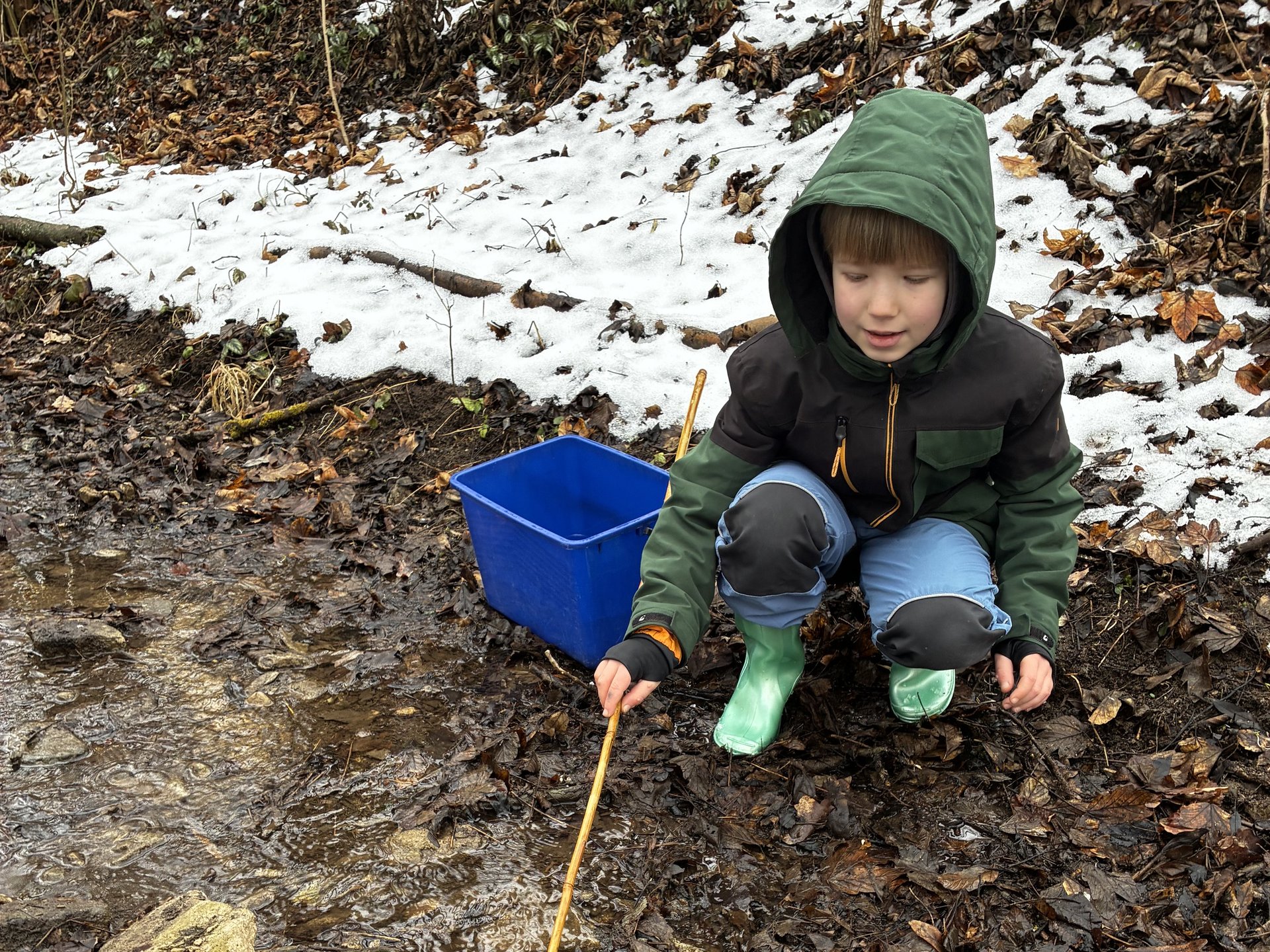 Ein Kind mit kniet am Ufer eines Bachs und hält ein Kescher ins Wasser; neben ihm steht ein blauer Eimer, im Hintergrund liegt Schnee.